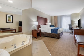 Quarto de hotel com uma cama grande, área de estar com um banco, escrivaninha, televisão e banheiro com banheira.