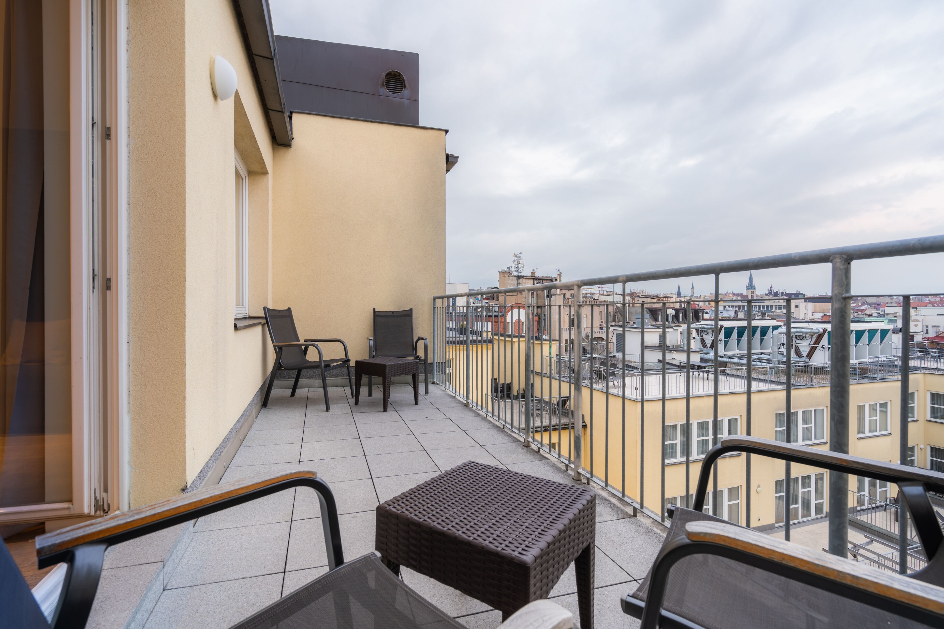 A balcony with a table and chairs, a small wicker ottoman, and a view of the cityscape.