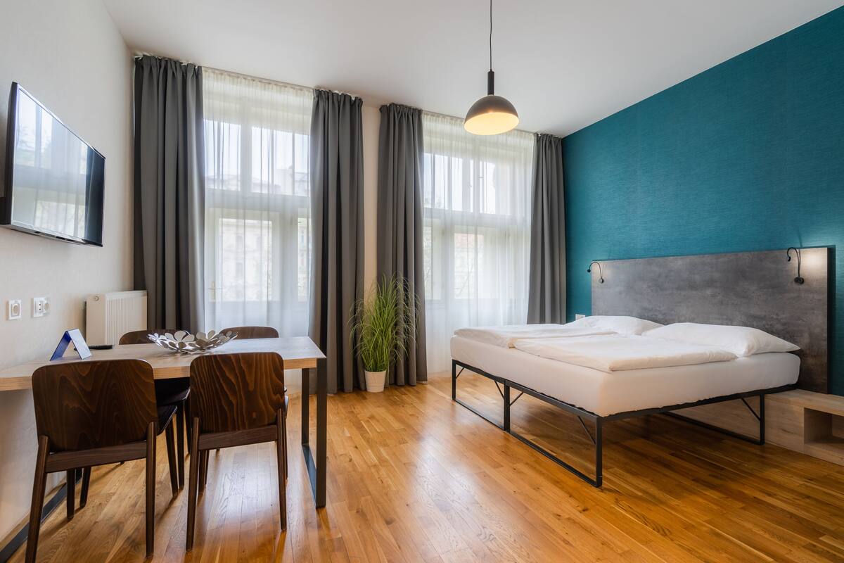 Une chambre d’hôtel moderne avec un plancher en bois, un lit, un bureau avec des chaises, une plante en pot et un téléviseur fixé au mur.