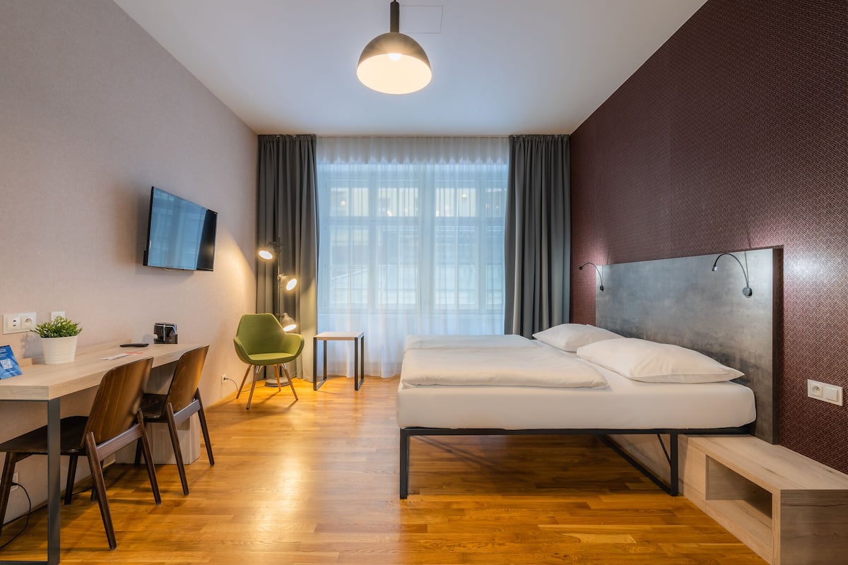 Une chambre d’hôtel moderne avec un plancher en bois, un lit avec du linge de maison blanc, un bureau avec des chaises, une télévision et une fenêtre avec des rideaux.