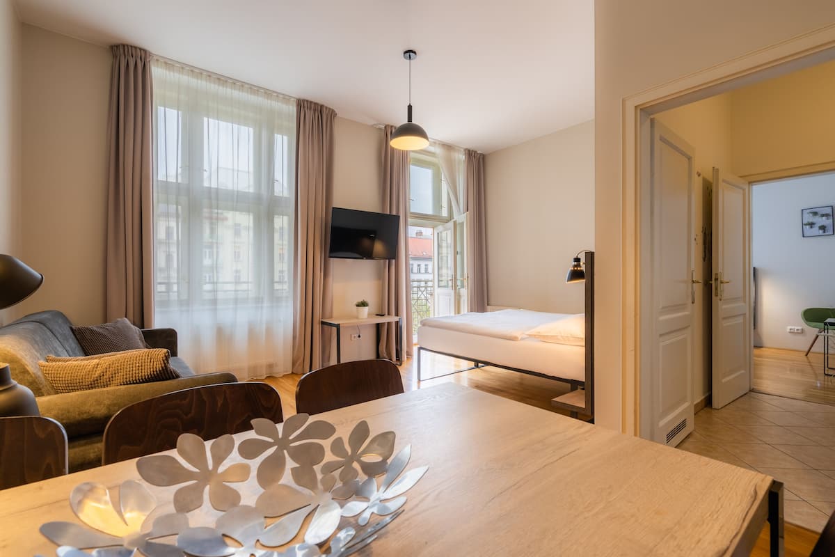 Une chambre d’hôtel comprenant un lit, un bureau avec une chaise, une table à manger agrémentée d’un élément décoratif, un téléviseur fixé au mur et de grandes fenêtres avec des rideaux.