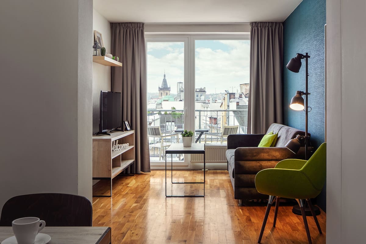 Un salon moderne avec un plancher en bois, un canapé, un fauteuil vert, un téléviseur et une vue sur le paysage urbain grâce à de grandes fenêtres.