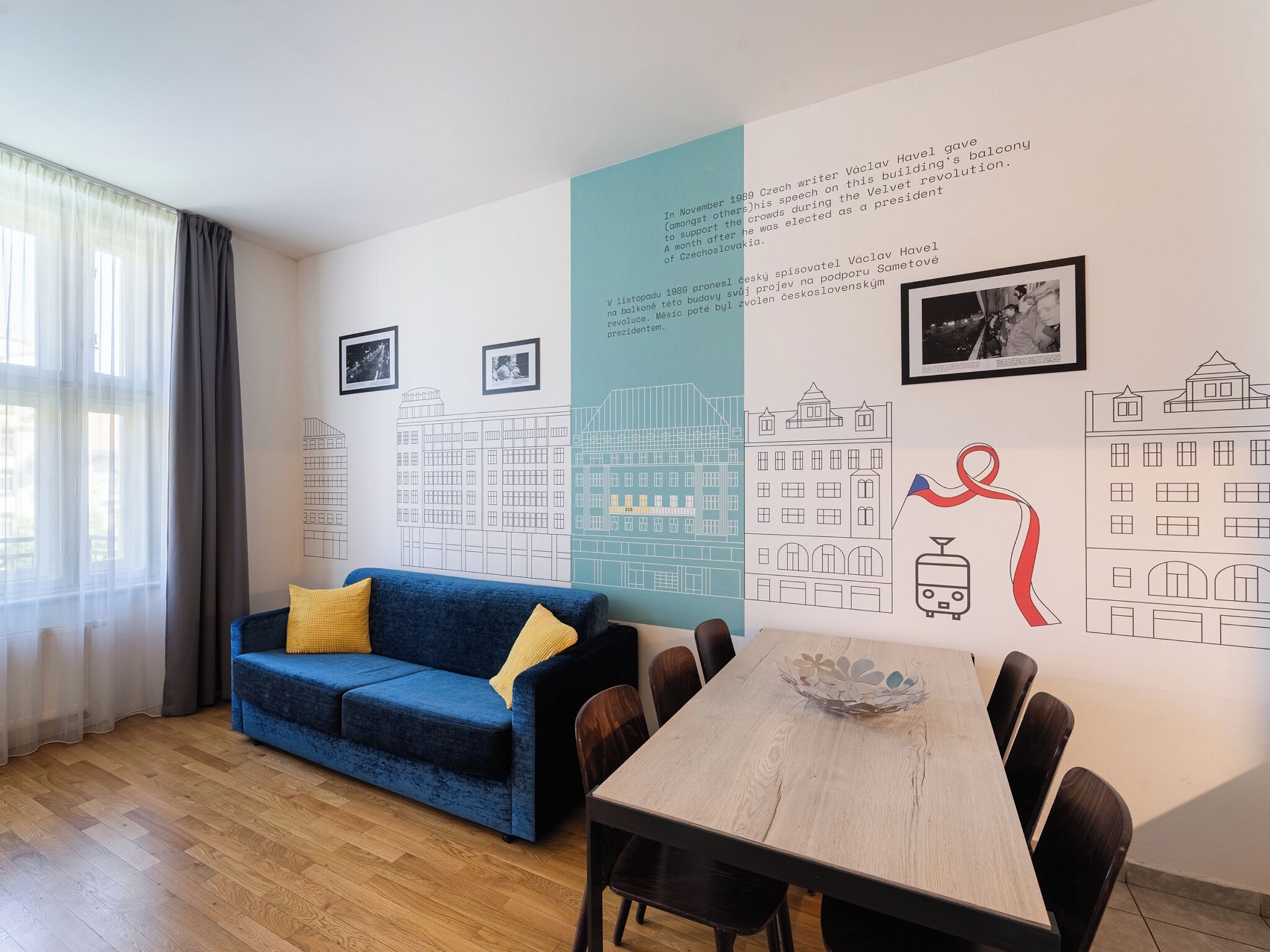 Una habitación moderna con un sofá azul, mesa de comedor de madera y sillas. Hay cuadros enmarcados en la pared y un mural grande que representa edificios y un símbolo de cinta.