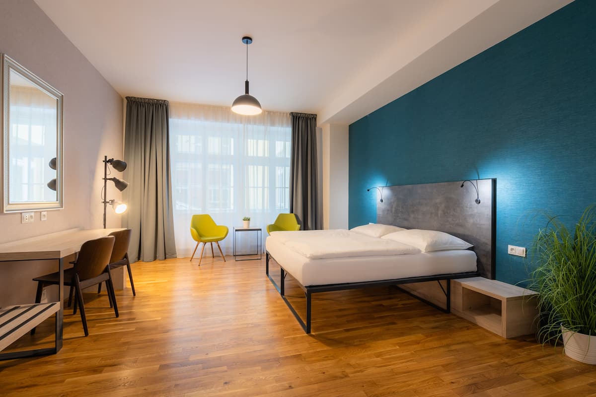 Une chambre d’hôtel moderne avec un plancher en bois, un lit, un bureau avec des chaises, une petite table et une plante en pot.