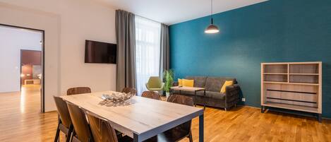 A modern living room with a dining area, wooden flooring, a blue accent wall, and a TV mounted on the wall.