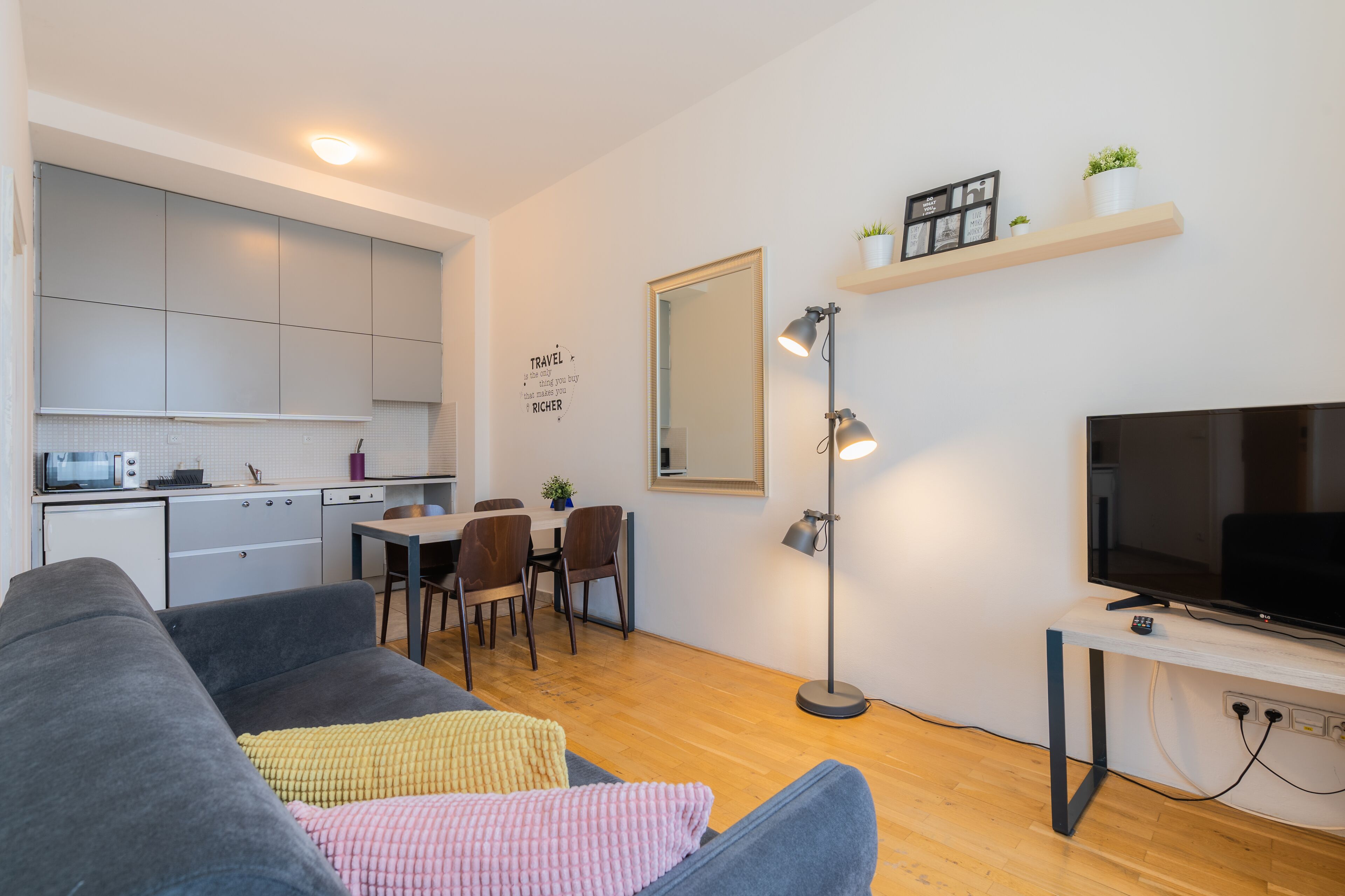 A modern living space with a grey sofa, a dining table with chairs, a kitchen area, a TV on a stand, and a floor lamp.