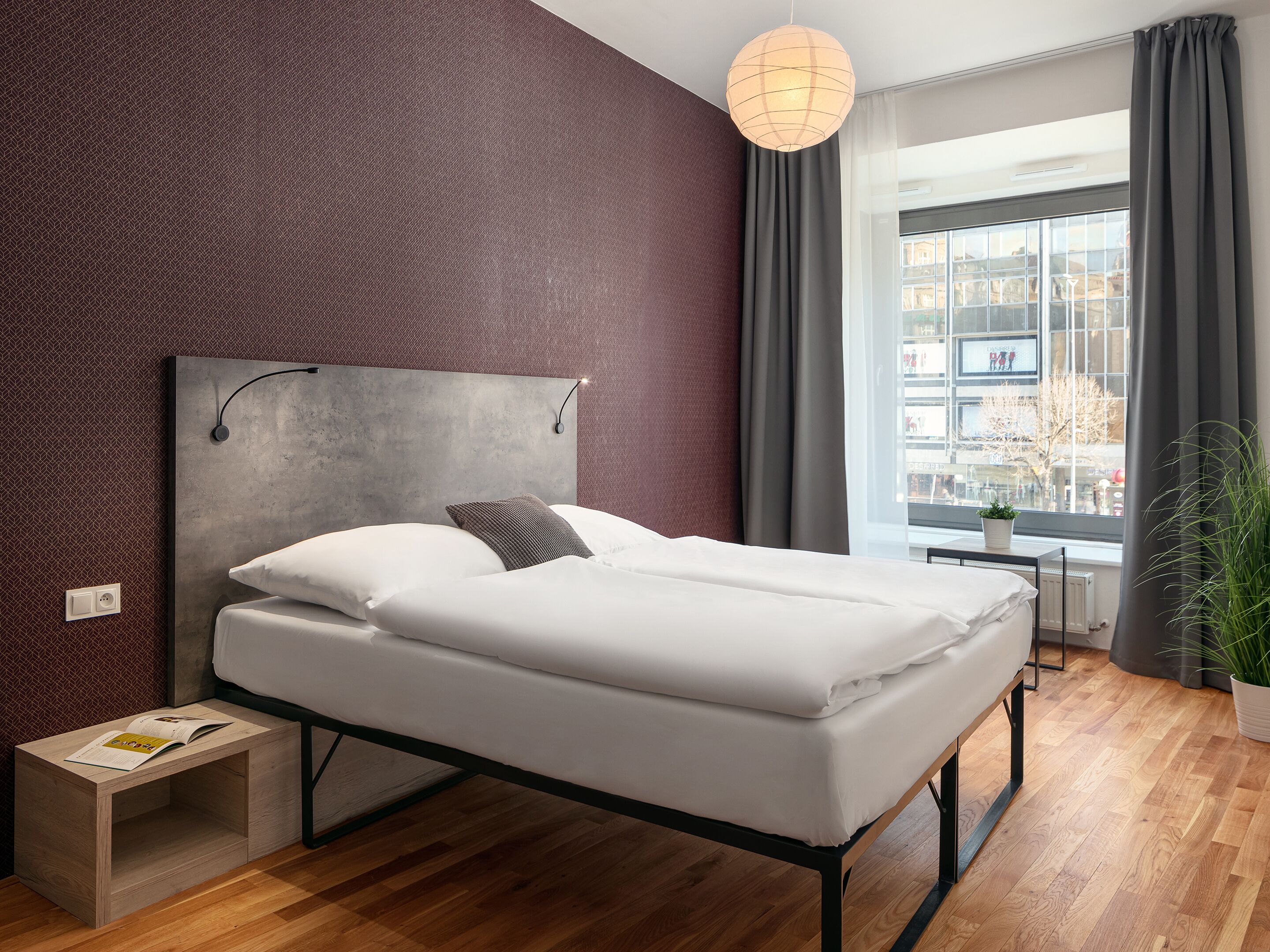 Een moderne slaapkamer met een groot bed, een nachtkastje, een hanglamp, een raam met gordijnen en uitzicht op gebouwen.