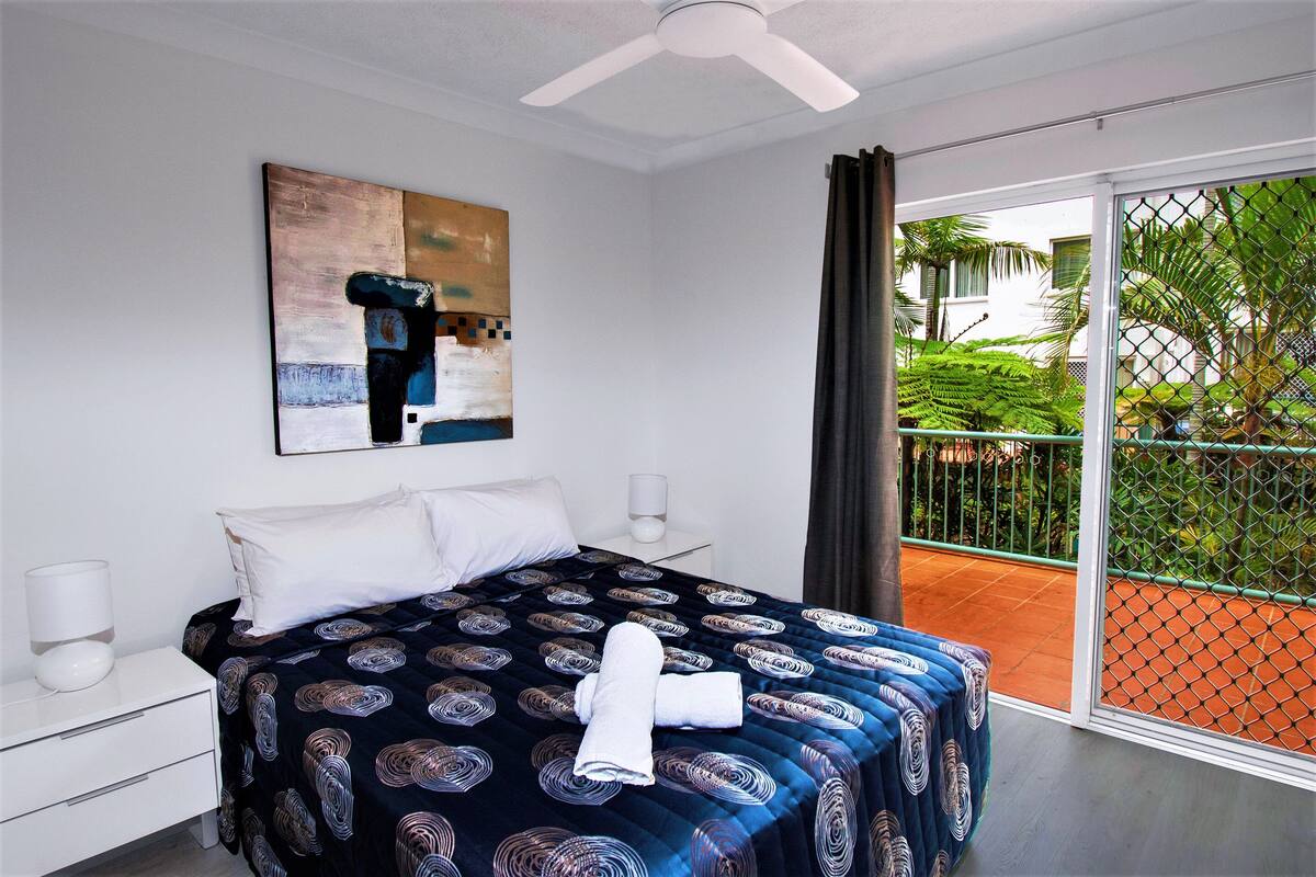 A hotel room with a bed, a ceiling fan, a painting, a window with a view of greenery, and a balcony with a fence.