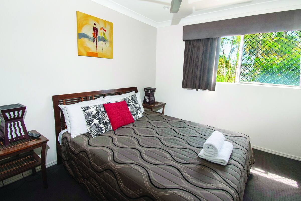 A neatly made bed with a patterned bedspread, a red pillow, and a rolled towel on the side. There is a small wooden nightstand with a lamp beside the bed. A painting hangs on the wall above the bed.