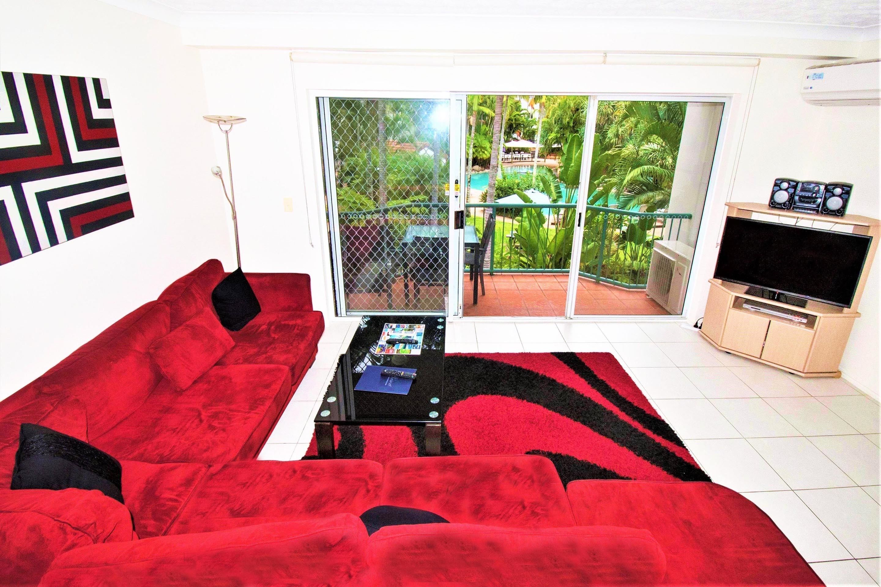 Una sala de estar con un sofá rojo, una mesa de centro, un televisor y un ventanal corredero que da a un balcón con vistas al verdor.
