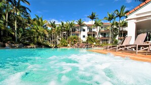 A hotel pool area with clear water, lounge chairs, palm trees, and a building with balconies.