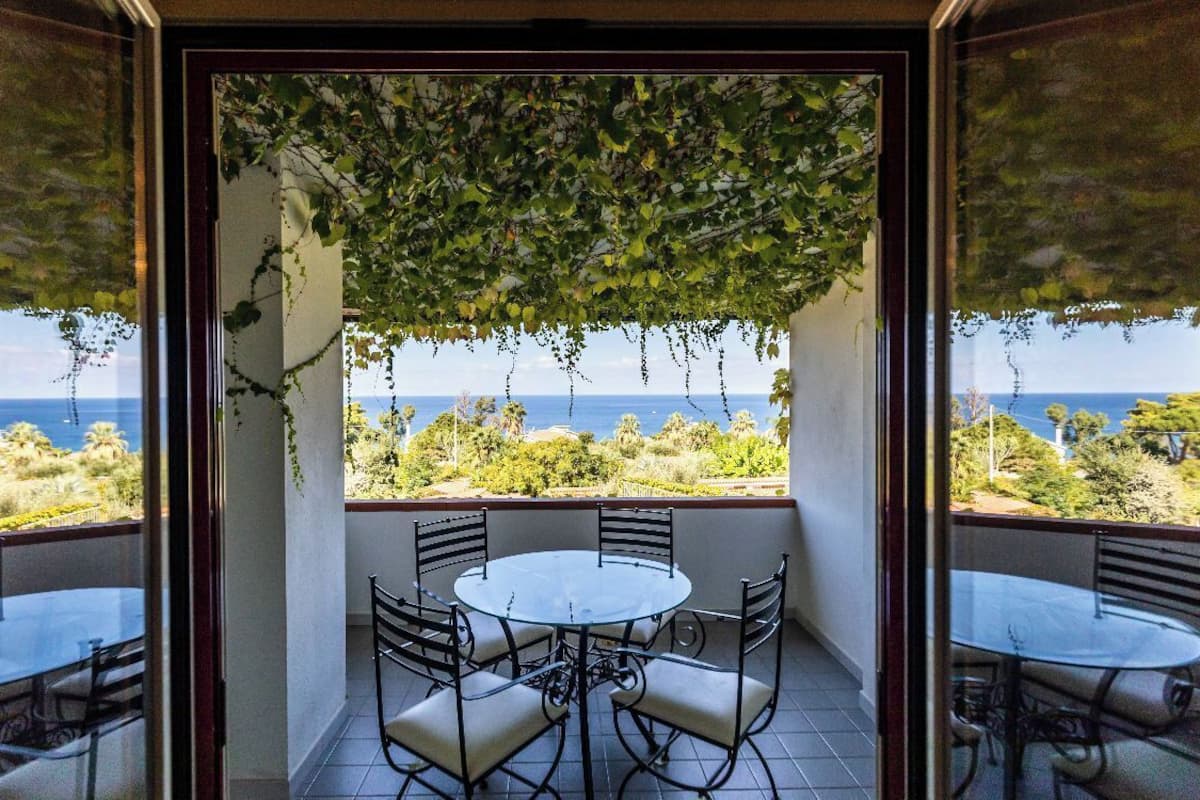 A balcony with a glass table and chairs, overlooking a scenic view of the sea and greenery.