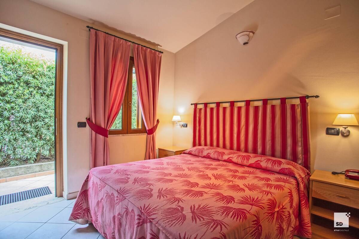 A bedroom with a bed, a window with curtains, a nightstand with a lamp, and a view of greenery outside.