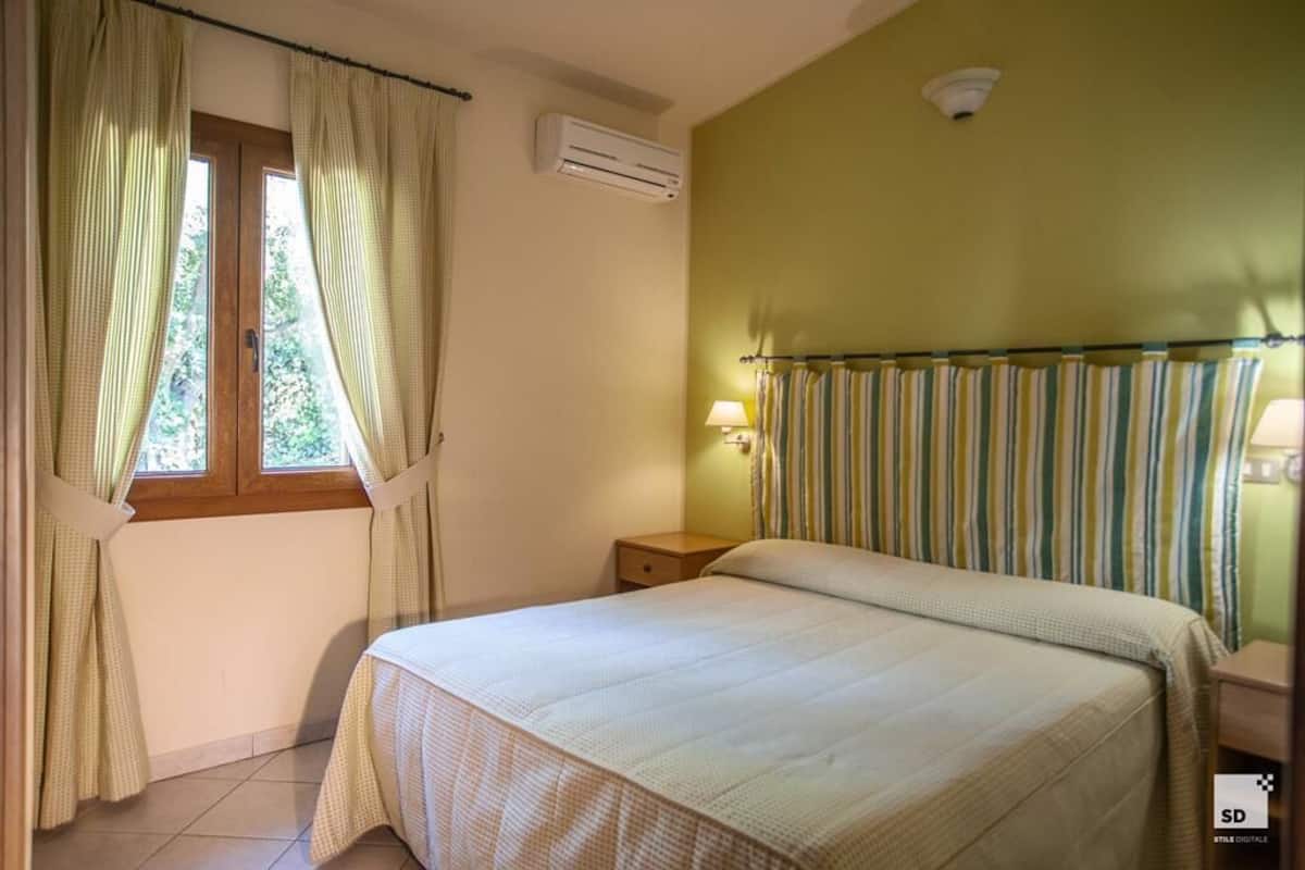 A bedroom with a bed, an air conditioner, a window with curtains, a bedside lamp, and a radiator.