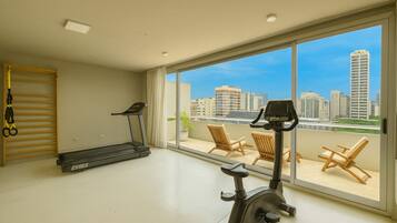 A spacious room with a treadmill and exercise bike, large windows with a city view, and a balcony with chairs.