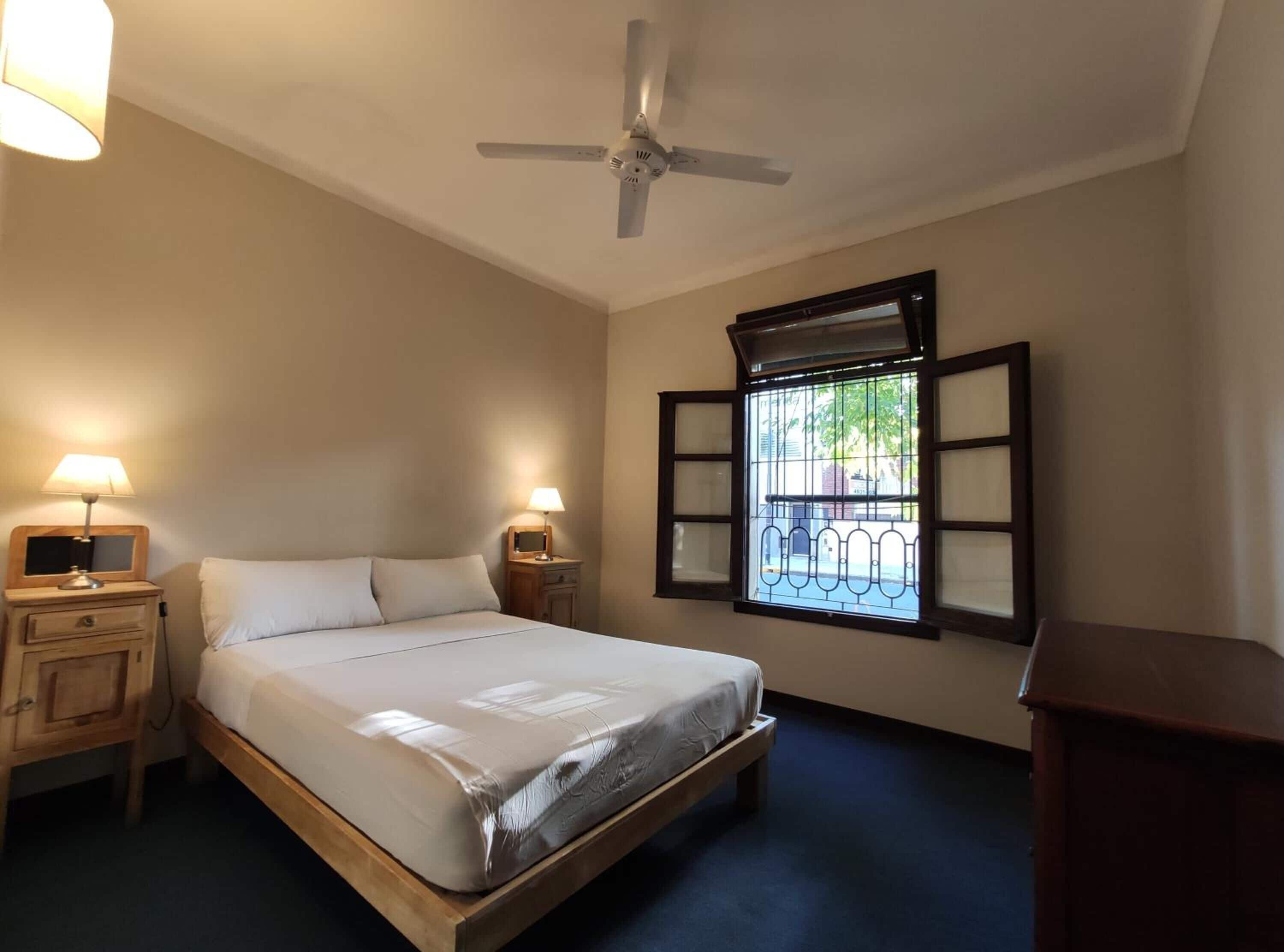 A bedroom with a single bed, two bedside tables with lamps, a ceiling fan, and a window with a metal grill.