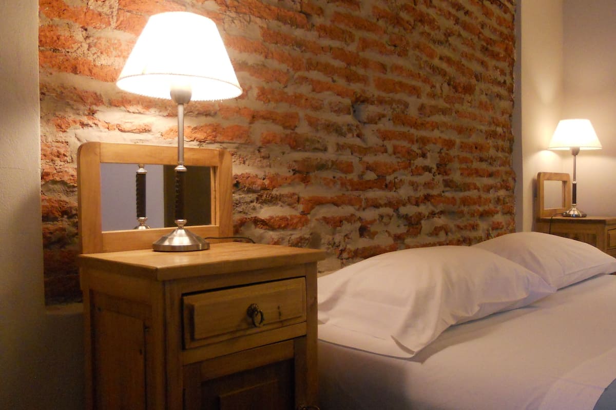 A hotel room with a brick wall, a wooden nightstand with a lamp, a mirror, and a bed with white linens.