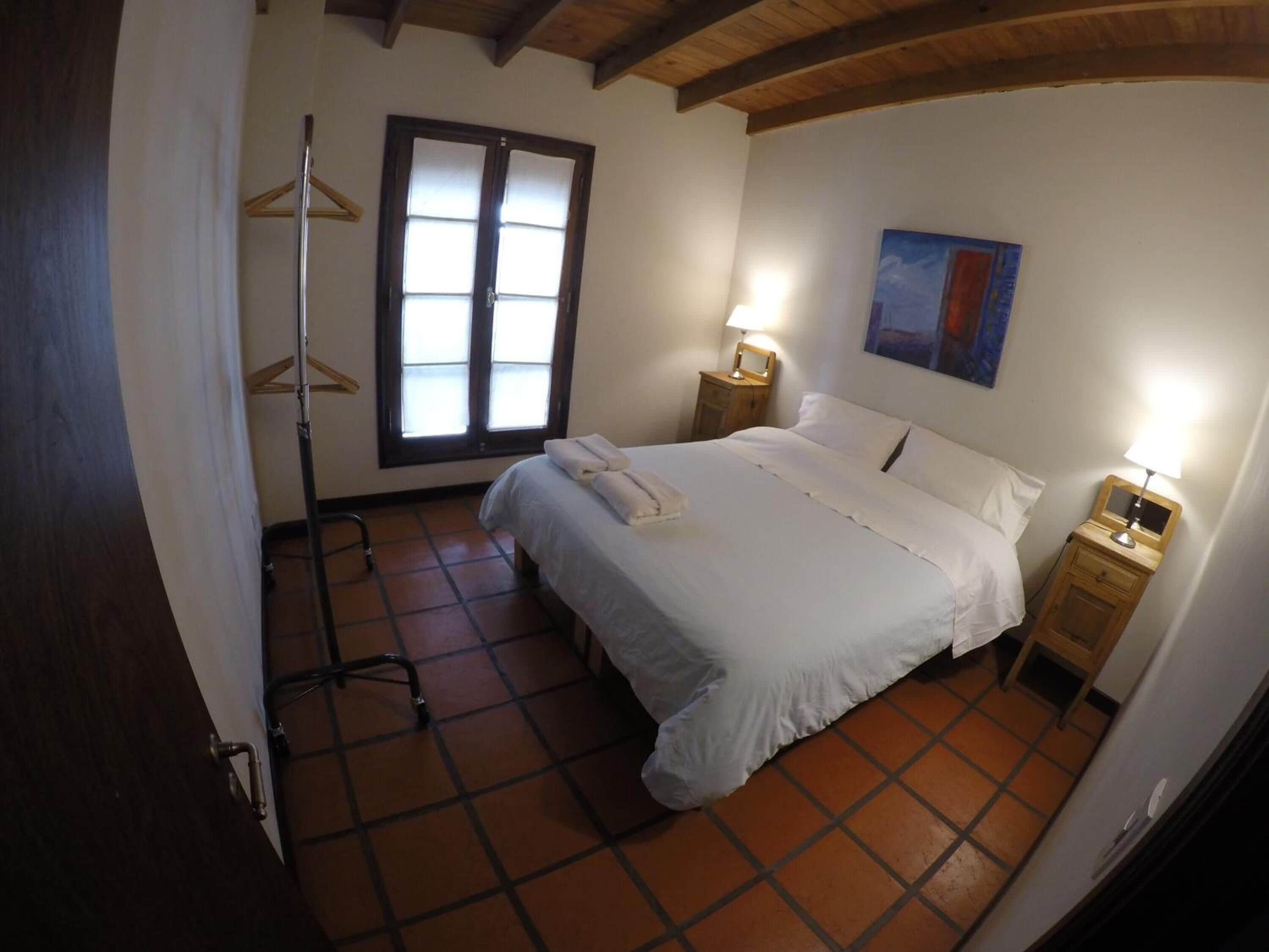 Un dormitorio con una cama, dos mesitas de noche con lámparas, una ventana con persianas, un perchero de madera y un cuadro en la pared.