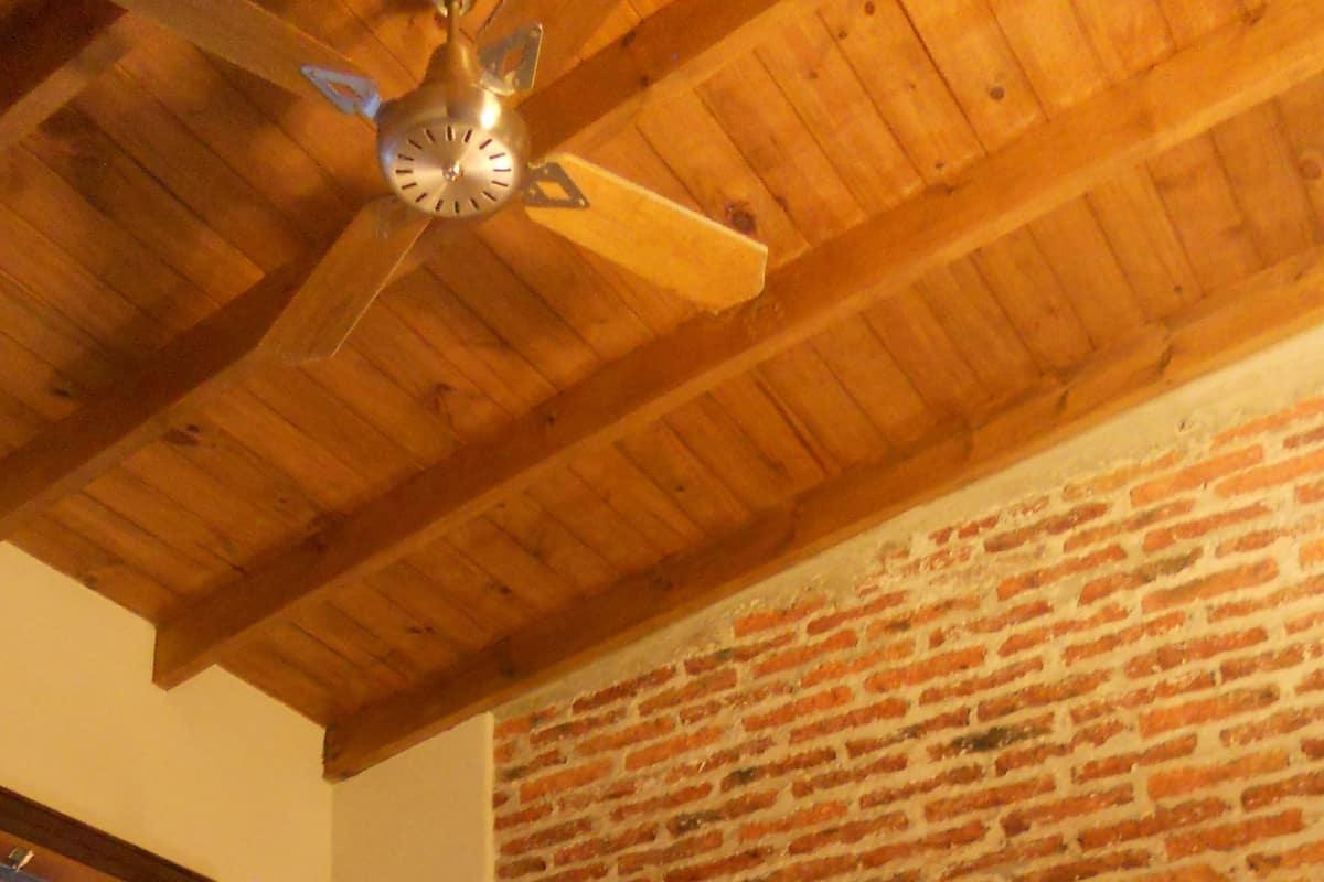 A room with a wooden ceiling, a brick wall, a bed with white linens, and a nightstand with a lamp.