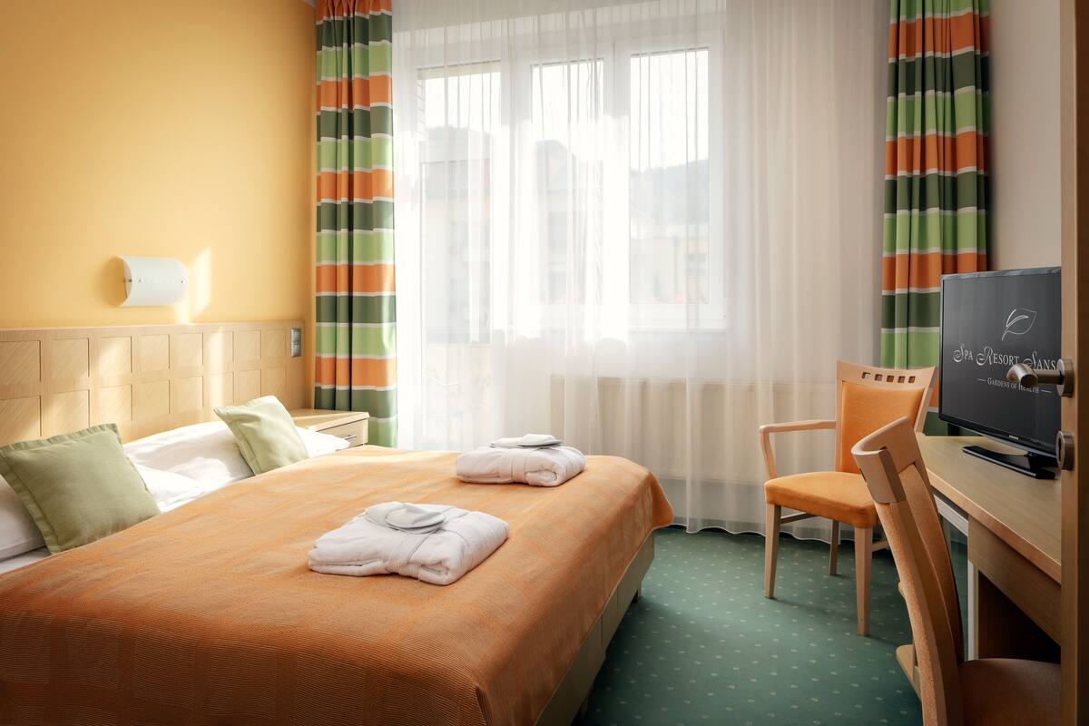Ein Hotelzimmer mit Bett, Sessel, Schreibtisch und Fernseher sowie einem Fenster mit Vorhängen.