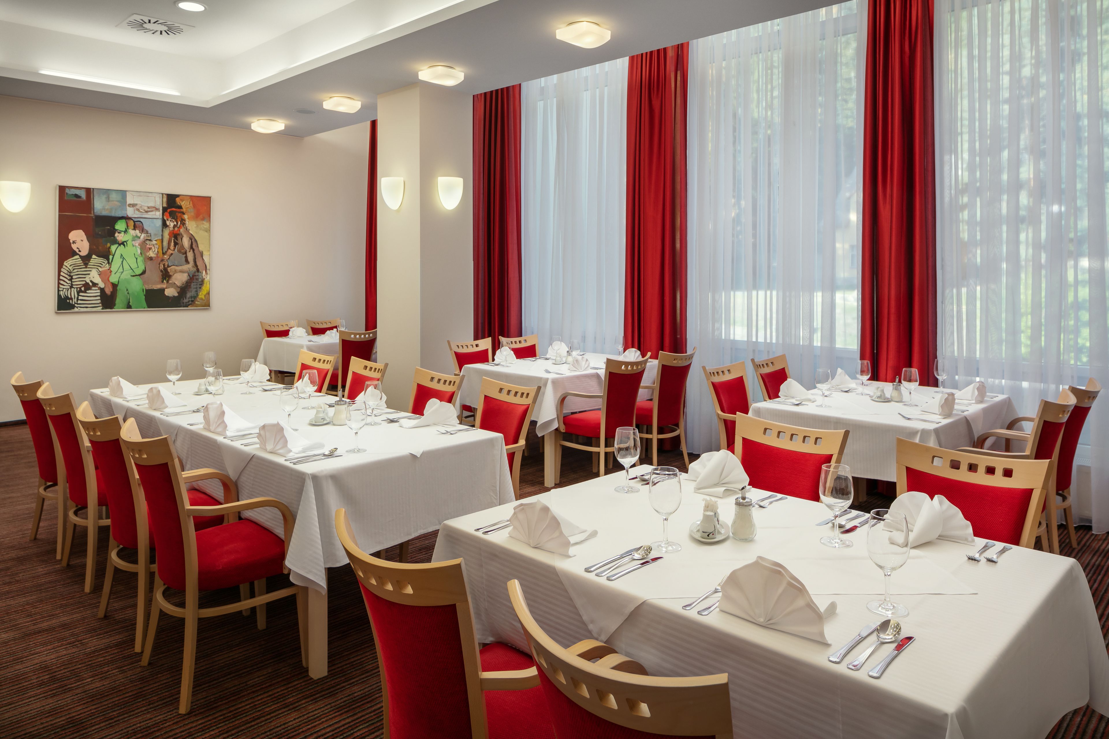 Un espace repas avec plusieurs tables dressées, des chaises à revêtement rouge et un tableau accroché au mur.