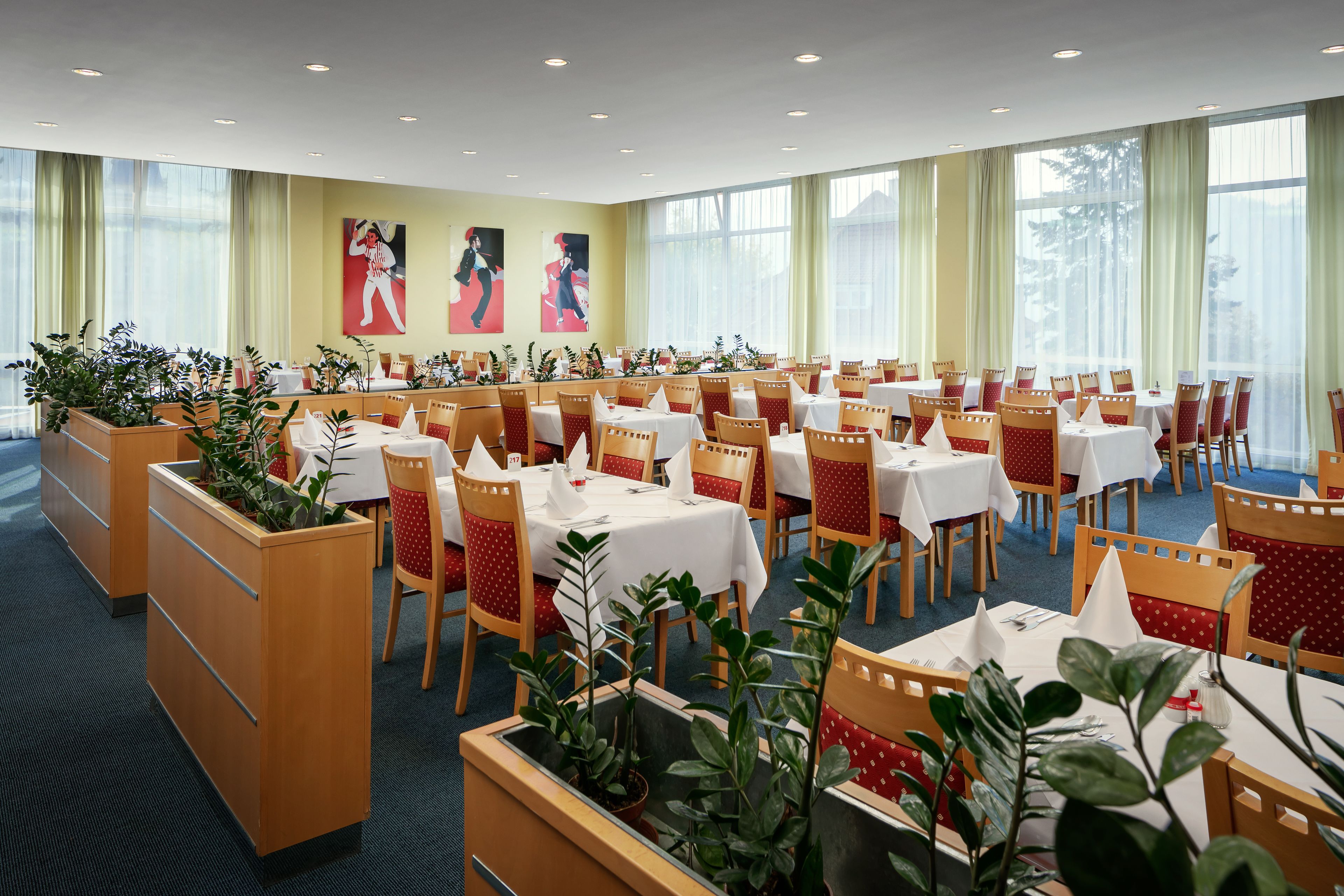 A hotel dining area with multiple tables and chairs, large windows, and indoor plants.