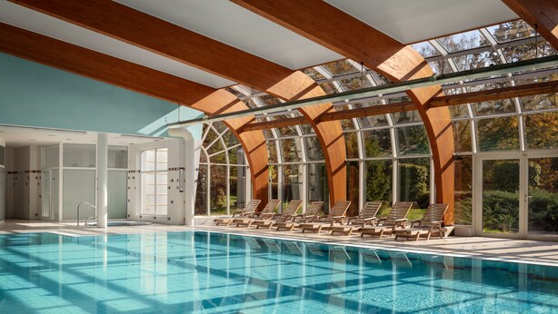Una piscina interior con techos de vidrio transparente y vigas de madera, rodeada de sillones de descanso.