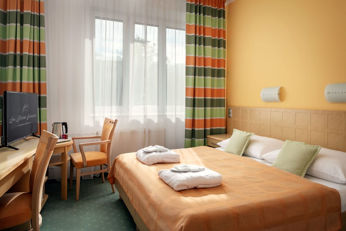 Ein Hotelzimmer mit einem Bett, einem Schreibtisch mit Computer, einem Stuhl und einem Fenster mit Vorhängen.