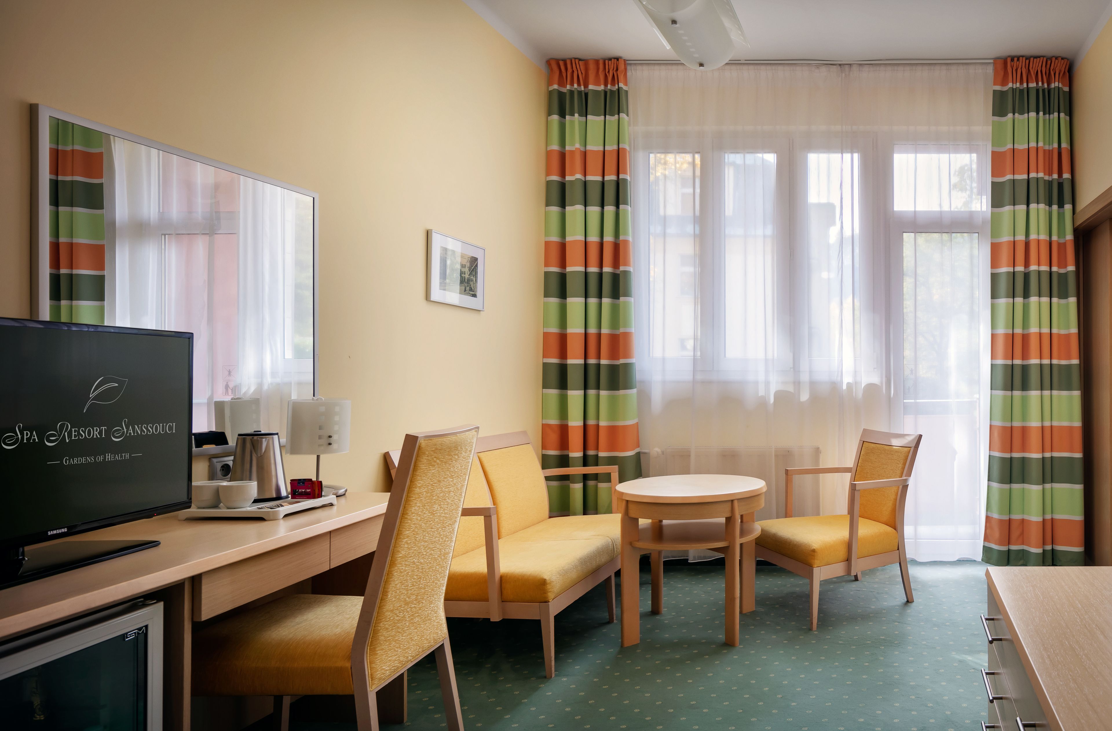 A hotel room with a TV, a desk, two chairs, a small table, and a window with curtains.