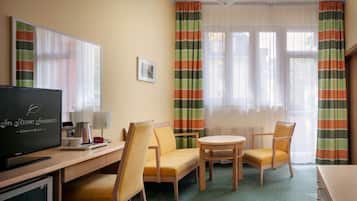 A hotel room with a TV, a desk, two chairs, a small table, and a window with curtains.