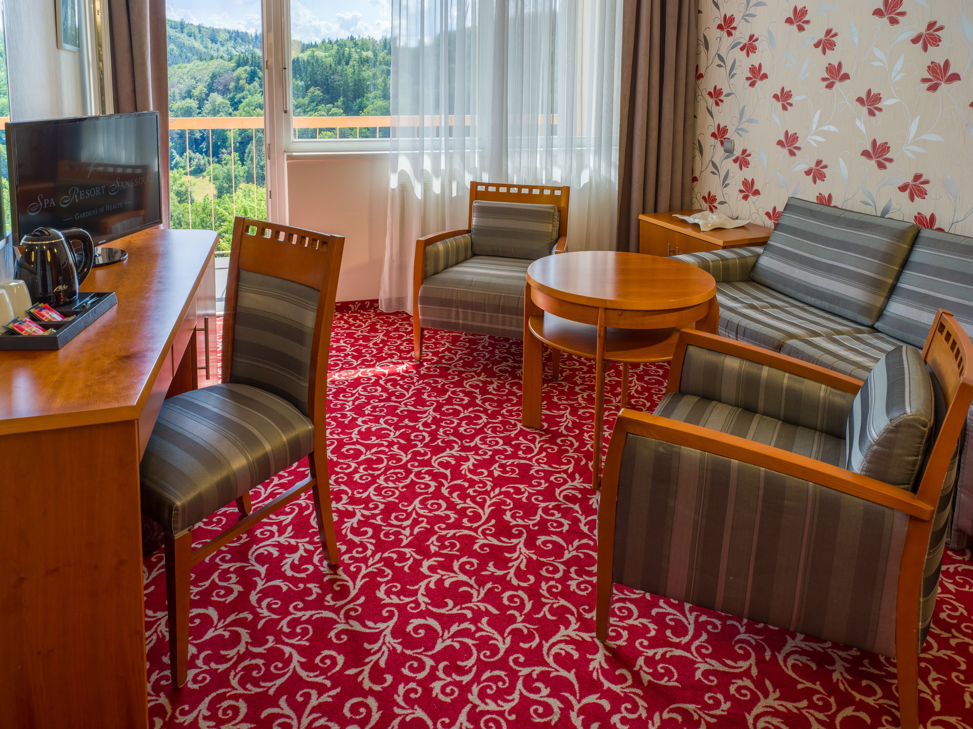 A hotel room with a red patterned carpet, a sofa, two armchairs, a small round table, and a television on a wooden stand.