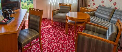 Una habitación de hotel con alfombra roja estampada, un sofá, dos sillones, una mesita redonda y un televisor sobre un mueble de madera.