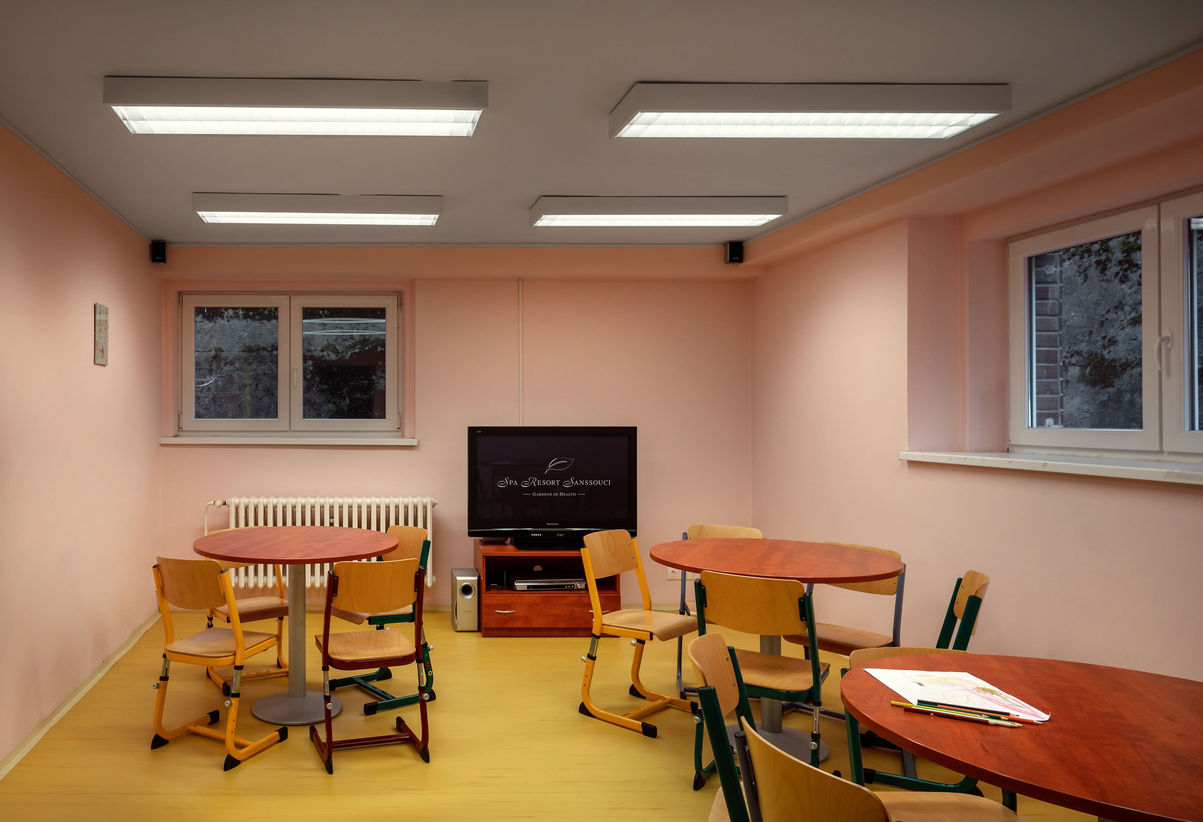 A conference room with a large flat-screen TV, multiple wooden tables and chairs, a radiator, and a window with a view of trees.