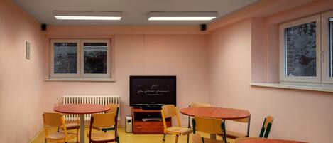 A conference room with a large flat-screen TV, multiple wooden tables and chairs, a radiator, and a window with a view of trees.