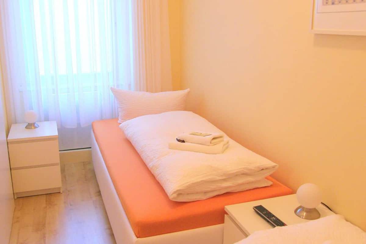 Un dormitorio pequeño y ordenado con una cama individual, una mesita de noche y una ventana con cortinas de encaje.