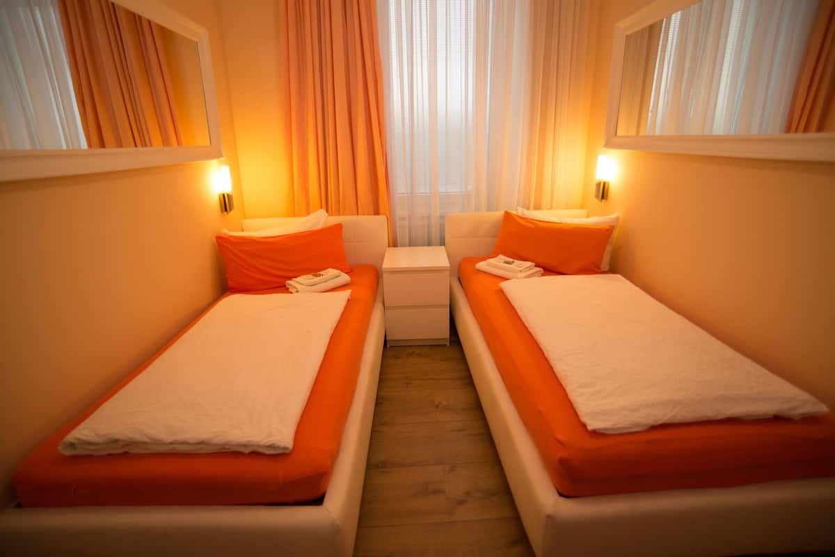 Habitación doble con dos camas, cada una con cubrecama naranja y almohadas blancas, una mesita de noche pequeña y una ventana con cortinas.