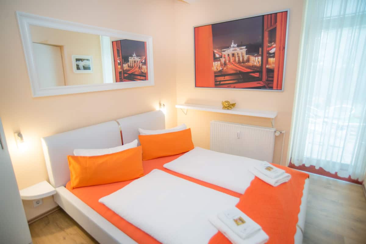 Habitación de hotel con cama, ropa de cama naranja, cabecero blanco, mesita de noche, espejo, cuadro enmarcado, estante con adorno y ventana con cortinas.