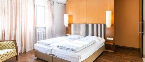 A modern hotel room with a large bed, wooden flooring, a sofa, and a window with curtains.