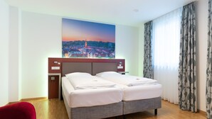 A hotel room with a bed, a red chair, a painting of a cityscape, and a window with curtains. - Altstadthotel Grauer Wolf (Erlangen)