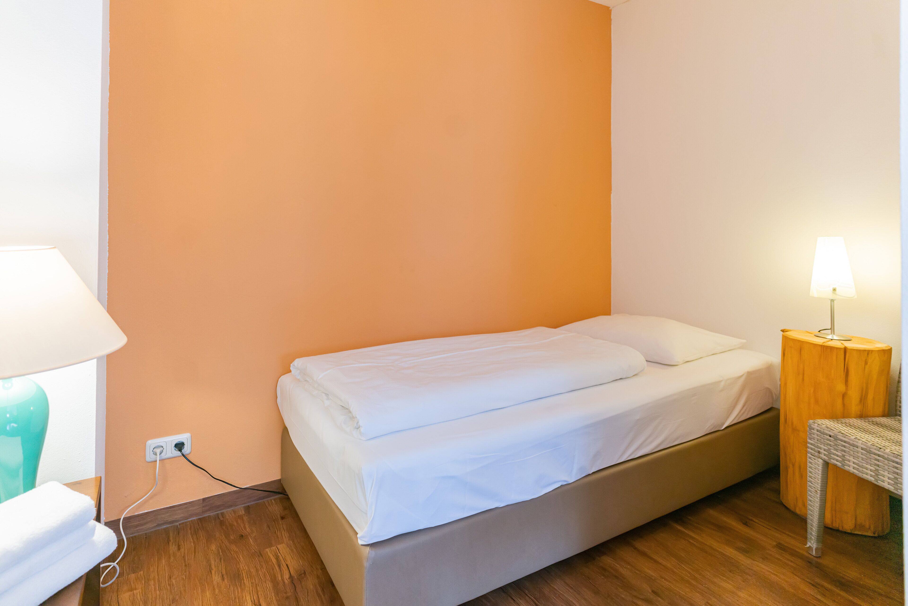 A single bed with white linens in a room with peach-colored walls and wooden flooring.