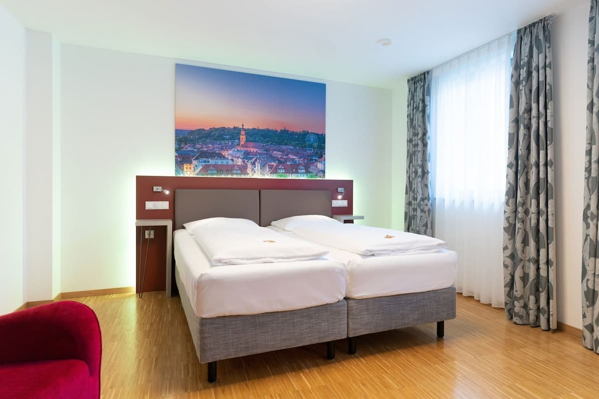 A hotel room with a double bed, a small desk, a chair, a large window with curtains, and a cityscape painting on the wall.