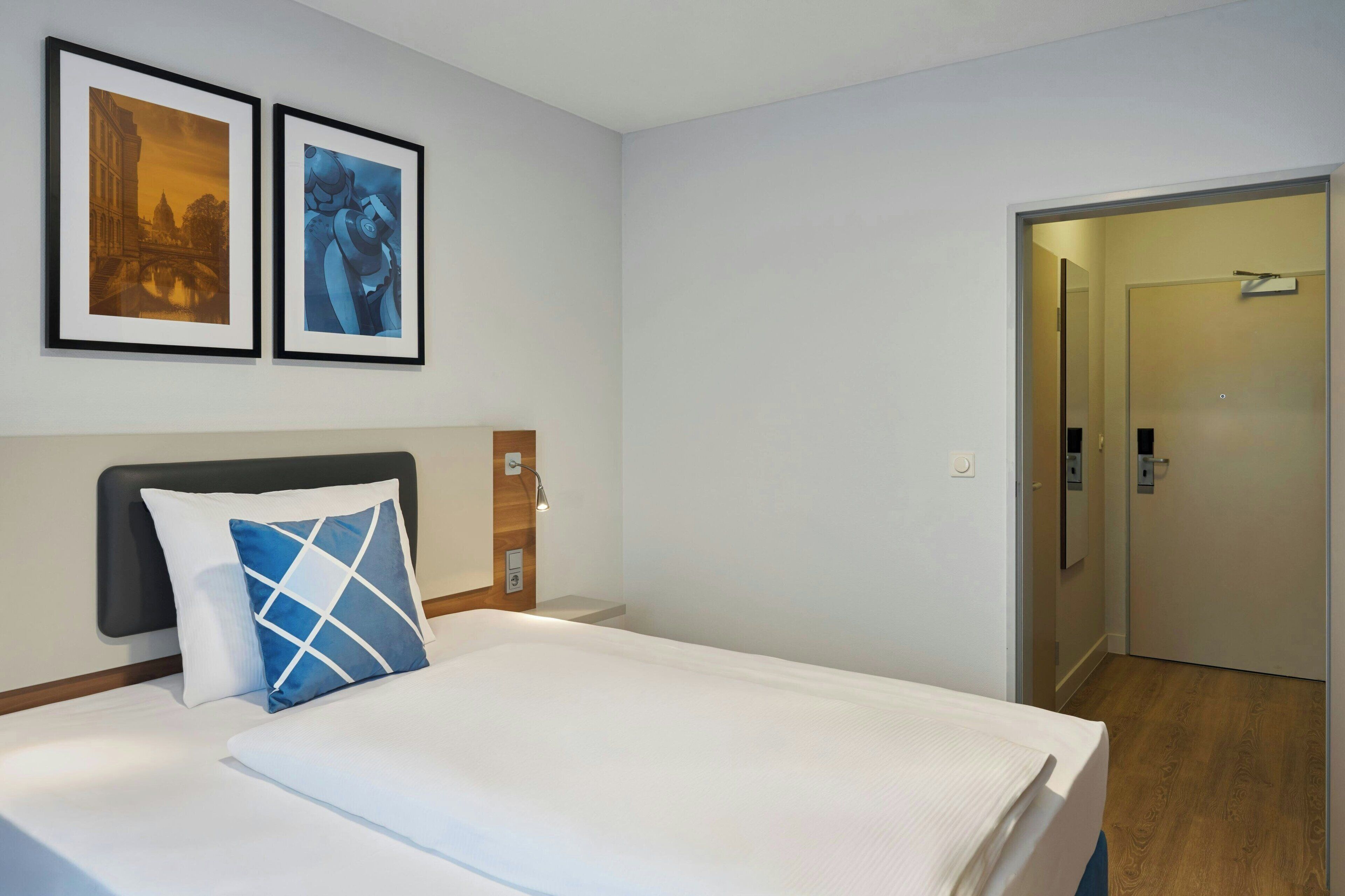 A hotel room with a bed, a headboard, two framed pictures on the wall, and a door leading to another room.