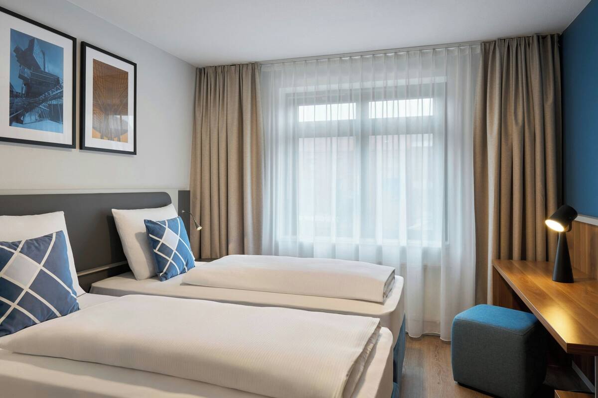 A hotel room with two beds, a desk, and a window with sheer curtains.