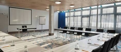 Una sala de conferencias con una gran mesa, sillas, una pantalla de proyector y un amplio ventanal con cortinas.