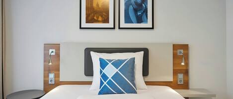 A hotel room with a bed, a nightstand, a wall-mounted lamp, and two framed pictures above the bed.
