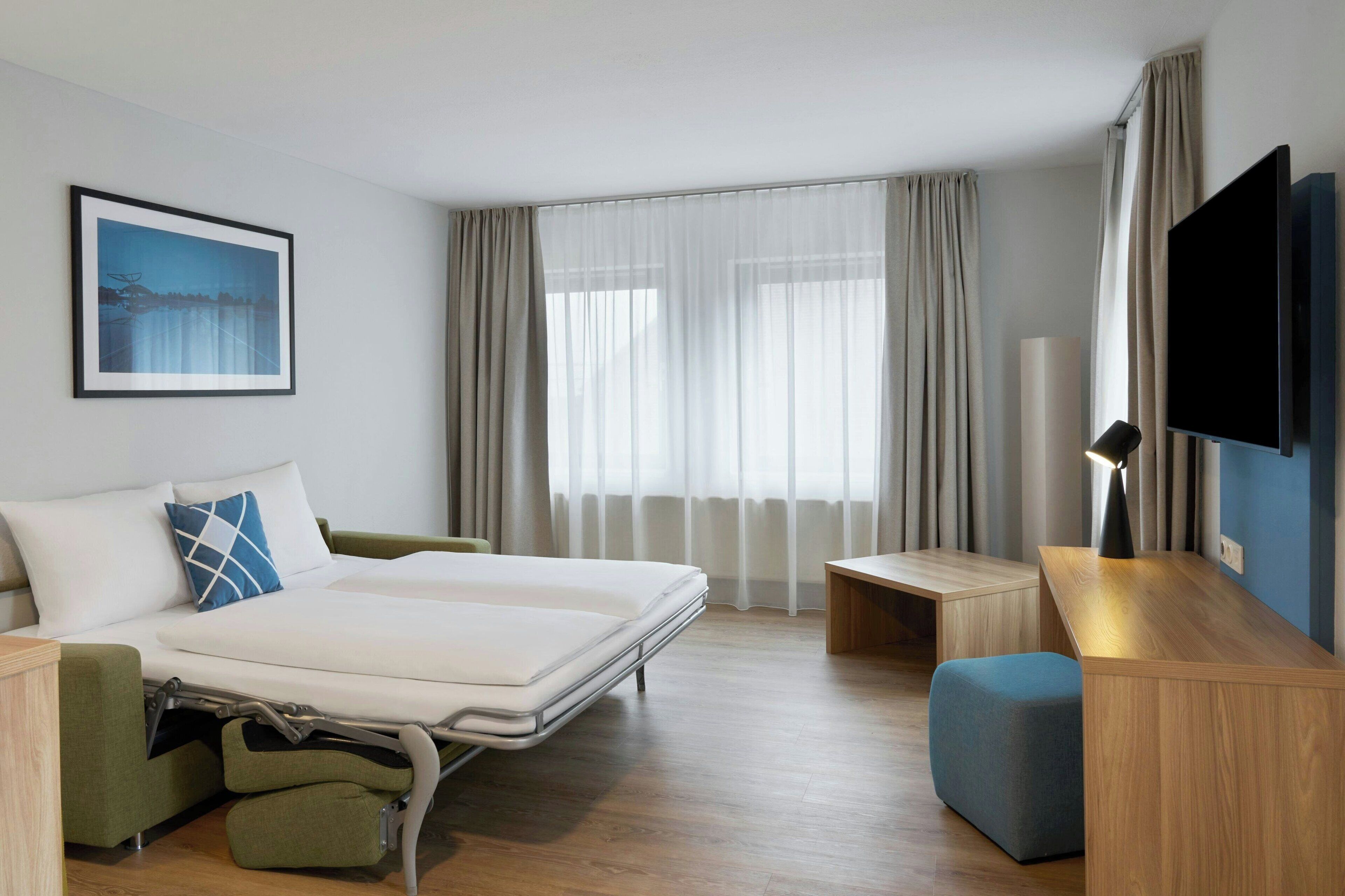 A modern hotel room with a bed, a desk, a TV, and a window with curtains.