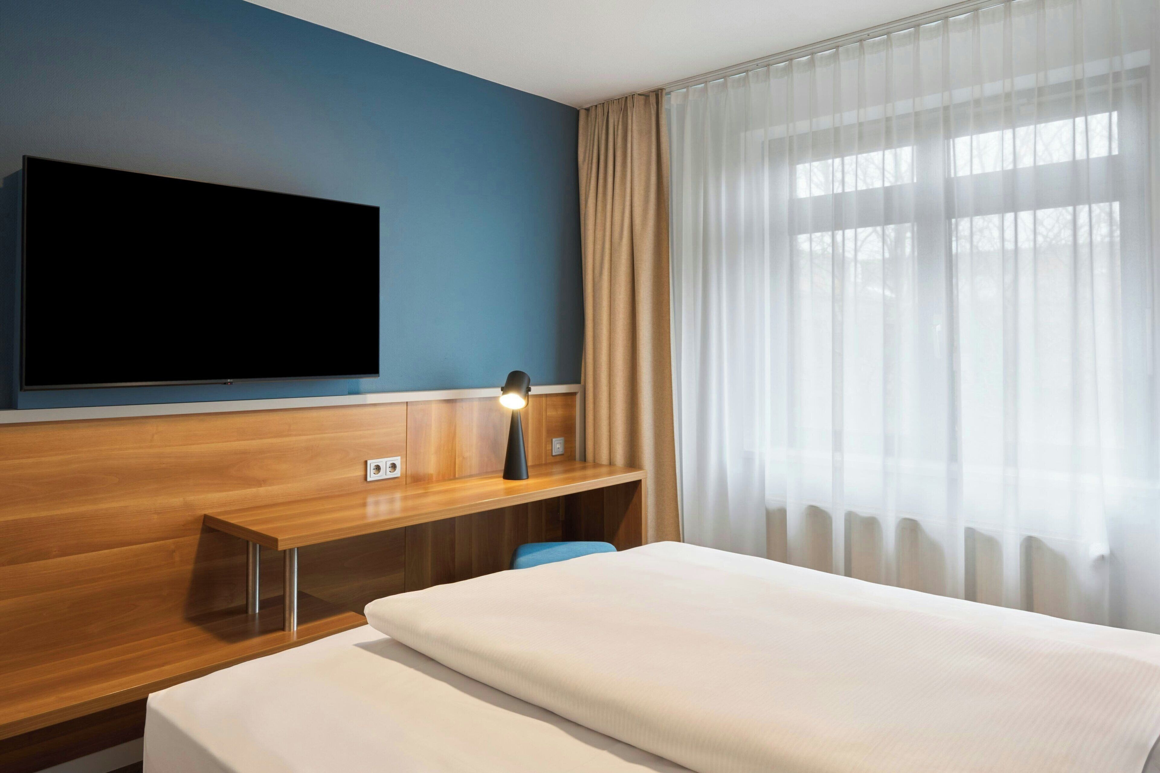 Une chambre d’hôtel avec un lit, un bureau doté d’une lampe, un téléviseur à écran plat fixé au mur et une grande fenêtre ornée de rideaux légers.