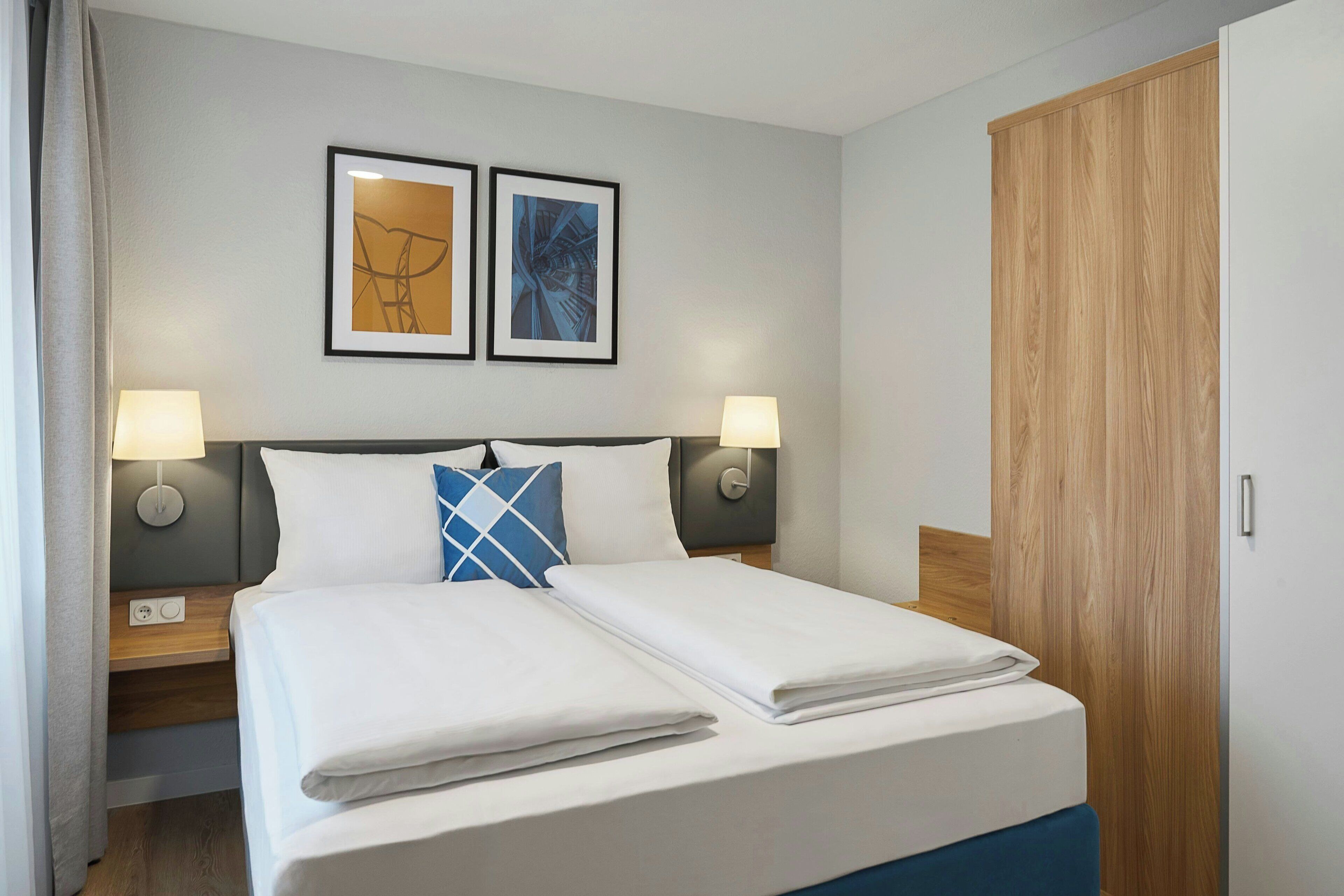 Habitación de hotel con cama doble, dos mesitas de noche con lámparas, un armario de madera y dos cuadros enmarcados en la pared.