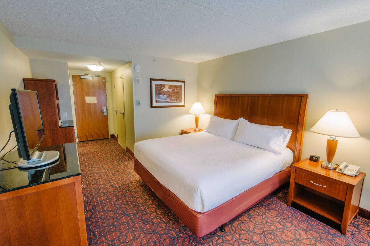 A hotel room with a large bed, two bedside tables with lamps, a desk with a computer, and a door leading to another room.