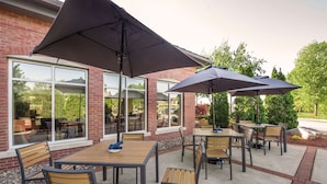 An outdoor patio with tables and chairs, large umbrellas, and a brick building with large windows. - Hilton Garden Inn Manchester Downtown (Manchester)